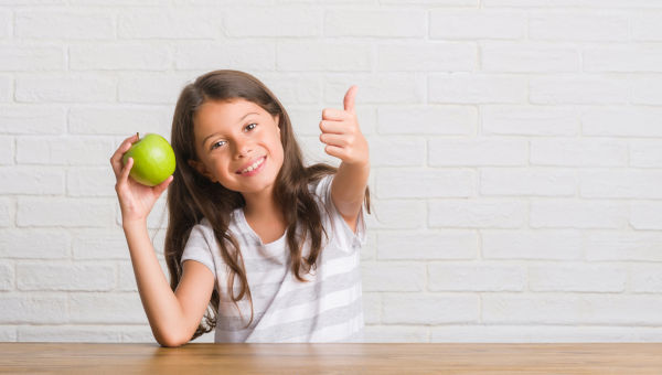 Junge, hispanische Kinder, die auf dem Tisch sitzen und frischen grünen Apfel essen glücklich mit großen Lächeln machen gutes Schild, Daumen mit den Fingern, ausgezeichnetes Schild