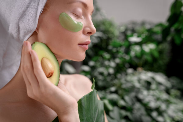 Charmantes Mädchen nach einer Dusche mit Patches unter den Augen posiert mit Avacado in den Händen auf unscharfem Hintergrund. Spa und Hautpflege