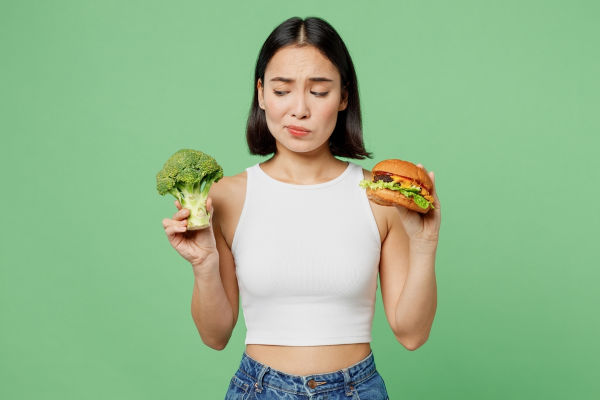 Junge Geistliche bedachte traurige Frau tragen weiße Kleidung halten Burger Broccoli Zähl Kalorien einzeln auf einfarbigem, hellgrünem Hintergrund. Richtige Ernährung gesunde Fast Food ungesunde Wahl Konzept