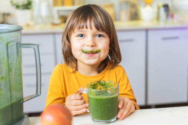 Happy Kid mit Glas Tasse grüner Smoothies in den Händen. Cute Junge verrückt trinkt gesunden diätetischen nährstoffreichen Cocktail zu Hause in der Küche. Gesunder Lebensstil, Rohkost