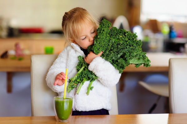 Klein Kleinkind in einer weißen, flauschigen Jacke hält einen Haufen frischen Kale und ist dabei, einen grünen Smoothie in der Küche zu trinken.